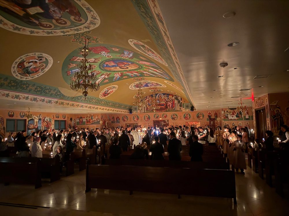 The inside of a Greek Orthodox church in Daly City at night, with painted walls and ceiling and a congregation holding candles