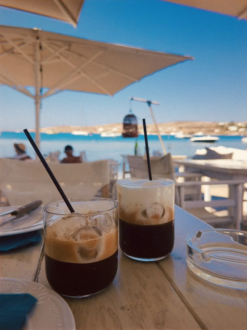 Two iced cappuccinos with black straws on a wooden table at a seaside café in Naxos, Greece, with white umbrellas and a blue bay behind