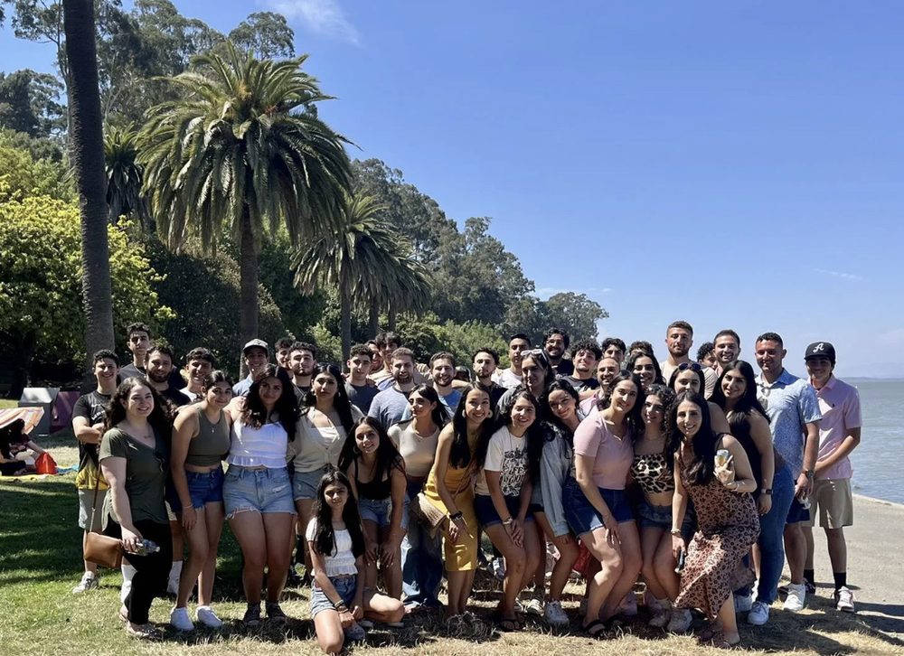A large group of Nabil's cousins posing together on a grassy shoreline with palm trees and water behind them