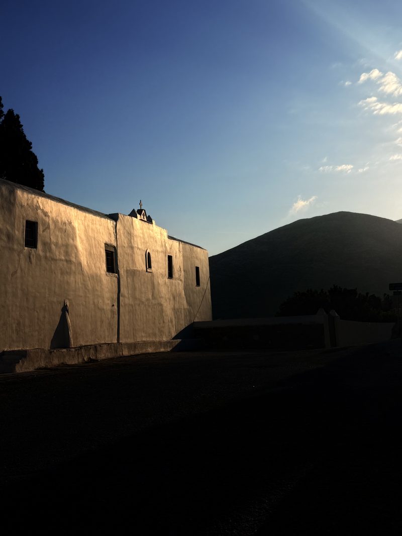 A whitewashed monastery with a small cross on its roof, lit by evening sun in front of a dark mountain